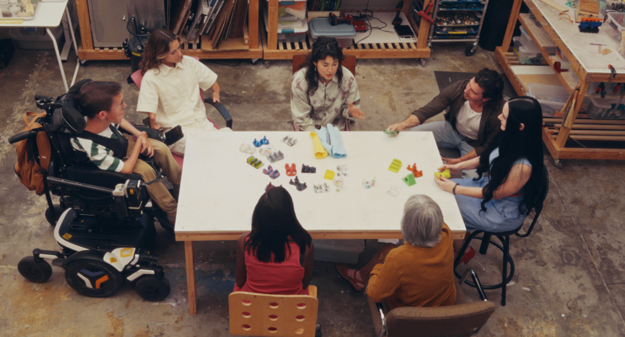 Un grupo de personas con diferentes necesidades de movilidad sentadas juntas en una mesa miran unos coloridos accesorios adaptativos para teléfonos inteligentes