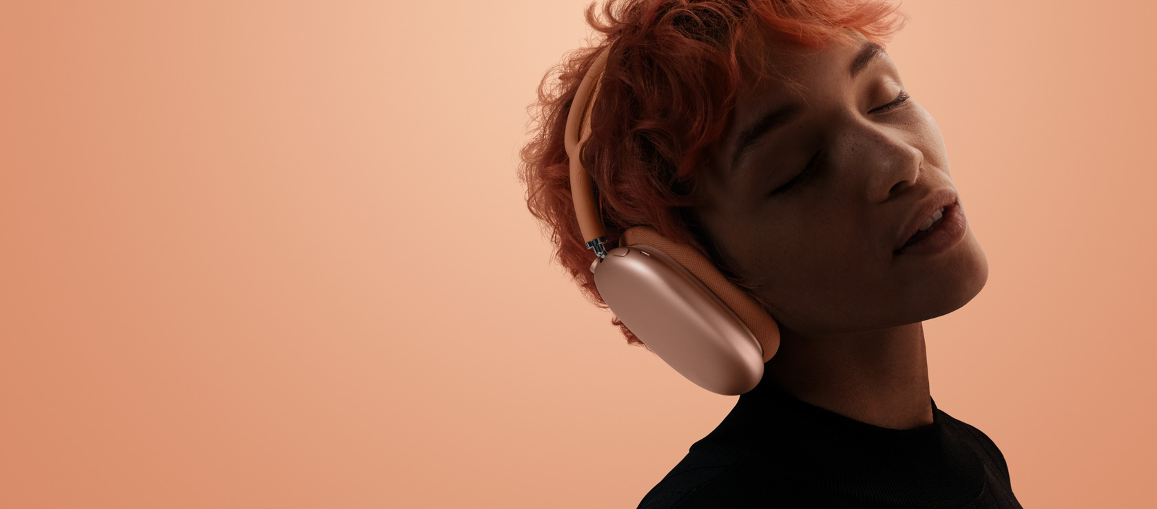 Človek so zavretými očami nakláňa hlavu. Má nasadené slúchadlá AirPods Max 2 oranžovej farby, oranžovo-ružové pozadie.