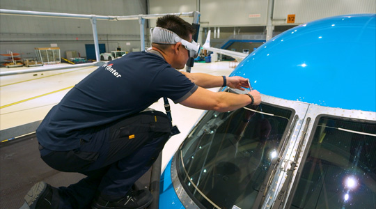 Une personne d’une équipe technique se trouve dans un hangar à avions; elle porte un Apple Vision Pro et tient un outil dans ses mains pour ajuster le pare-brise d’un avion