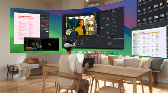 Une personne portant un Apple Vision Pro est assise à un bureau avec un MacBook Pro 13 pouces ouvert, le contenu de l’écran du Mac est projeté dans la pièce dans un affichage incurvé très large