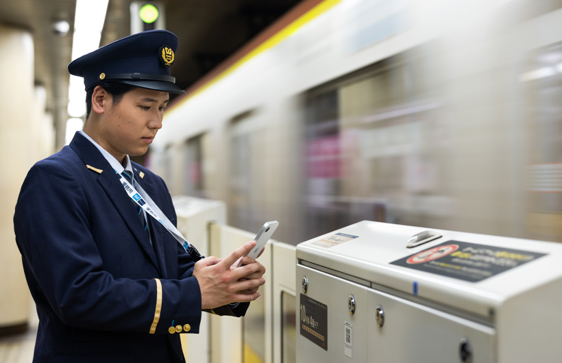 En stasjonsbetjent står på plattformen og bruker iPhone til å skanne en QR-kode mens en T-bane kjører forbi.