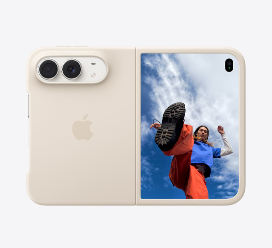 iPhone Duo, finition blanc stellaire, dans une coque en silicone sable, mode paysage, entièrement ouvert à l’horizontale, double caméra Fusion en haut à gauche, une coque recouvre la face extérieure arrière et le bord externe de l’écran avant, le côté droit présente l’écran externe affichant l’image d’une personne debout sous un ciel bleu nuageux